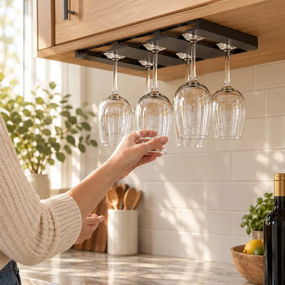 A woman in a cream knit sweater reaches up to slide a glass into a black under-cabinet wine glass rack mounted beneath a light wood cabinet, where four stemmed glasses already hang upside down above a sunlit marble counter with a wine bottle and a bowl of lemons nearby.